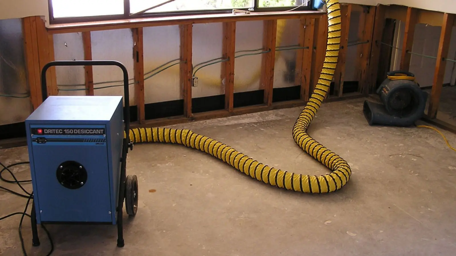 Dehumidifier with a yellow duct hose running in a room where the lower wall panels have been opened to dry